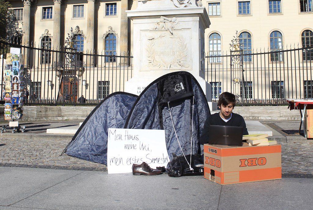 Ein Mann sitzt mit einem Karton, Zelt und Laptop vor einem Gebäude